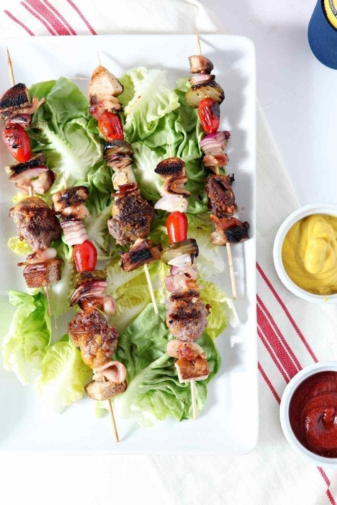 Overhead image of several Cheeseburger Kebabs, laid out on top of lettuce on a white platter, served with ketchup and mustard