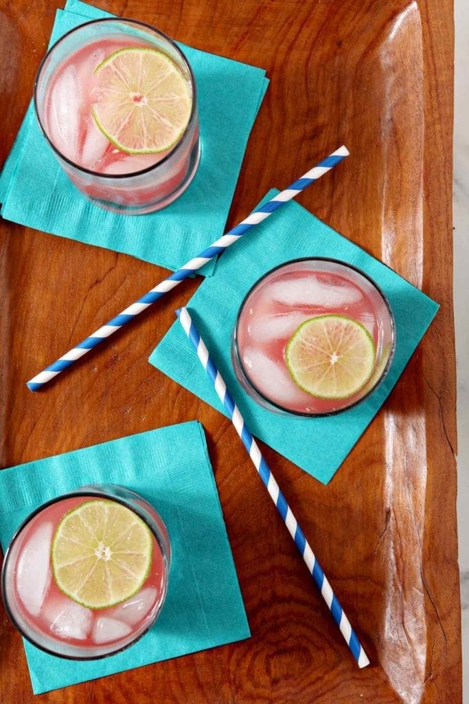 Three glasses of Spiked Watermelon Limeade sit on a wooden serving platter, served on turquoise napkins with blue striped straws