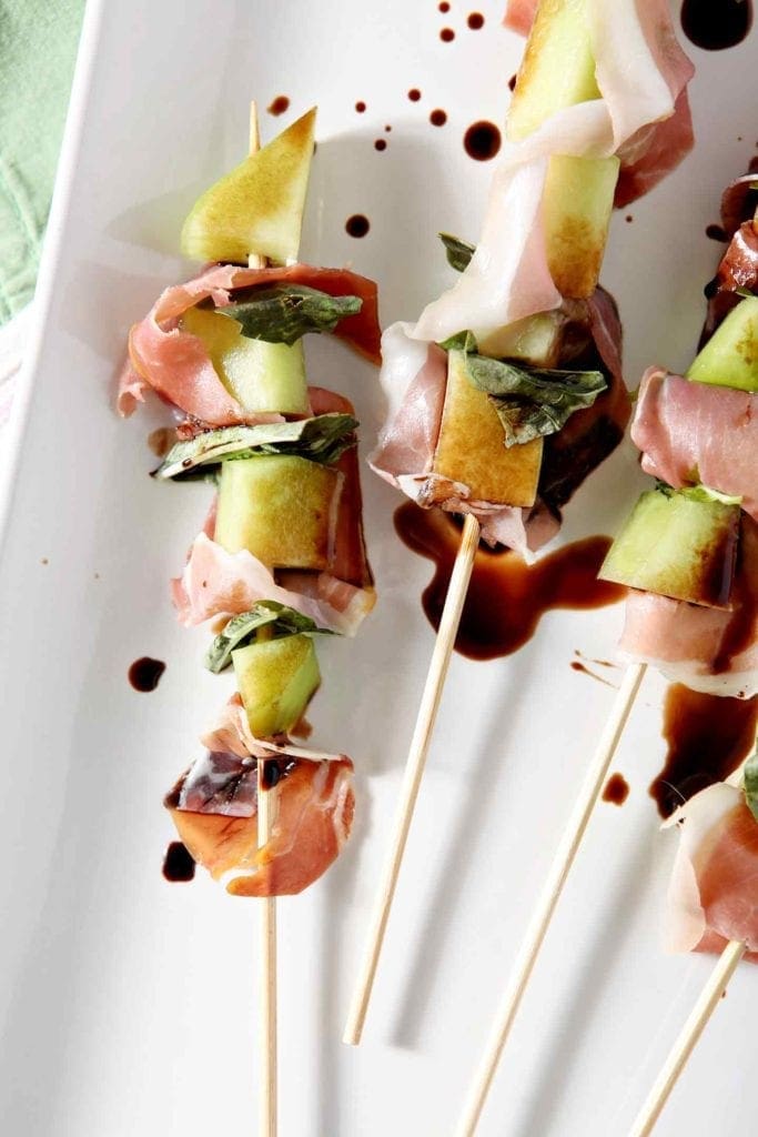 Close up of Galia Melon Jamón Skewers (Whole30 Appetizer) on a white platter, drizzled with balsamic vinegar, before serving.