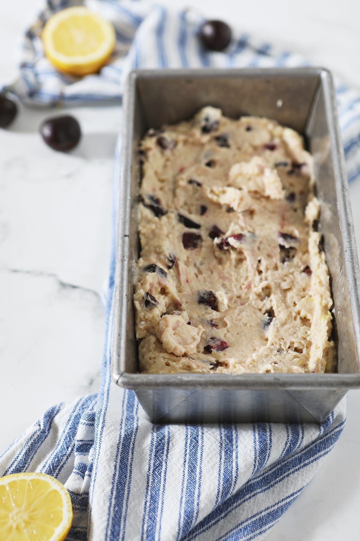 Cherry Bread batter in a metal bread pan on top of a blue and white striped towel before baking