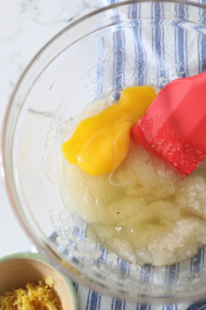 A broken egg on top of a sugar mixture in a clear glass bowl with a red rubber spatula