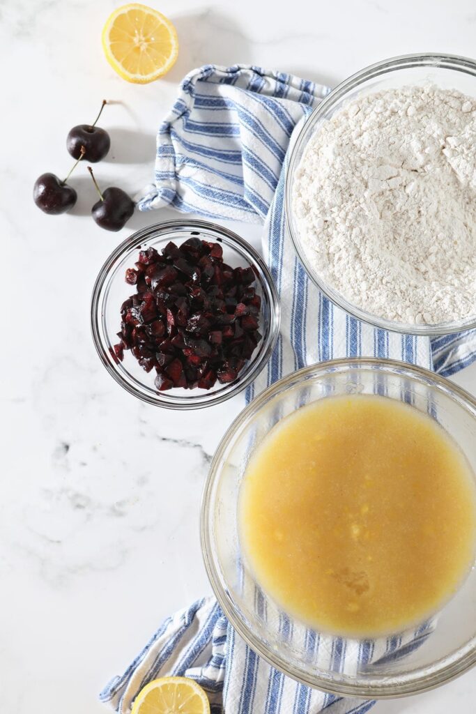 Overhead of wet ingredients, dry ingredients and chopped cherries on top of a blue striped towel