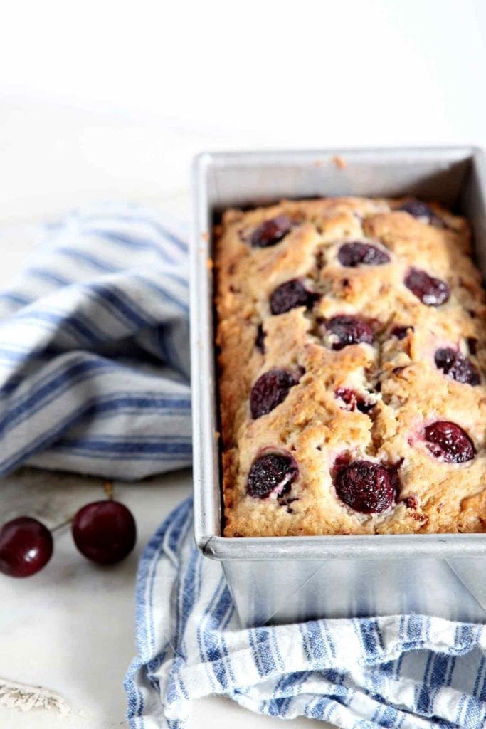 A loaf of Fresh Cherry Bread in its pan on a blue and white striped towel, next to fresh cherries