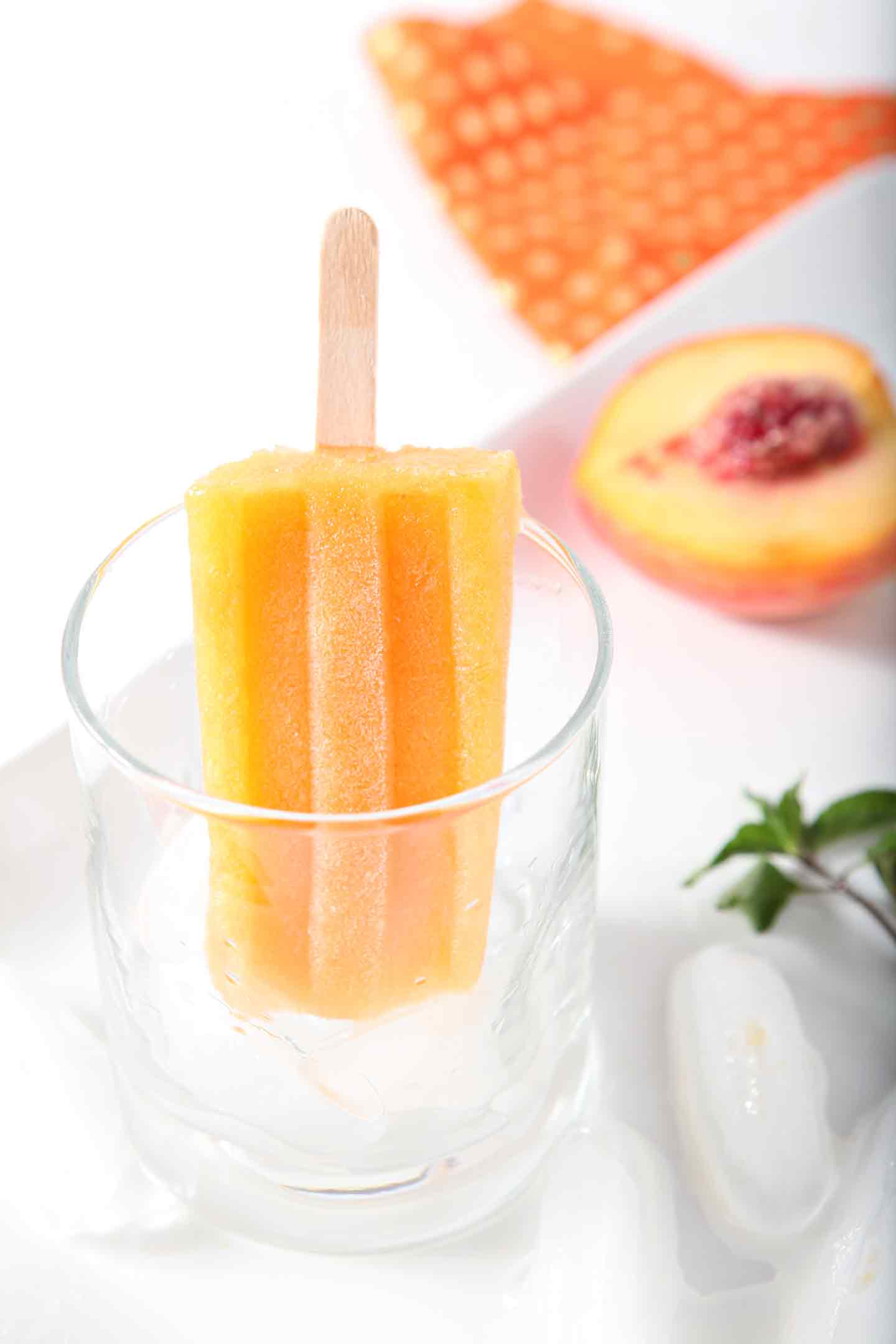 A lone Peach Julep Popsicle hangs out in a glass, surrounded by a halved peach and sprigs of mint