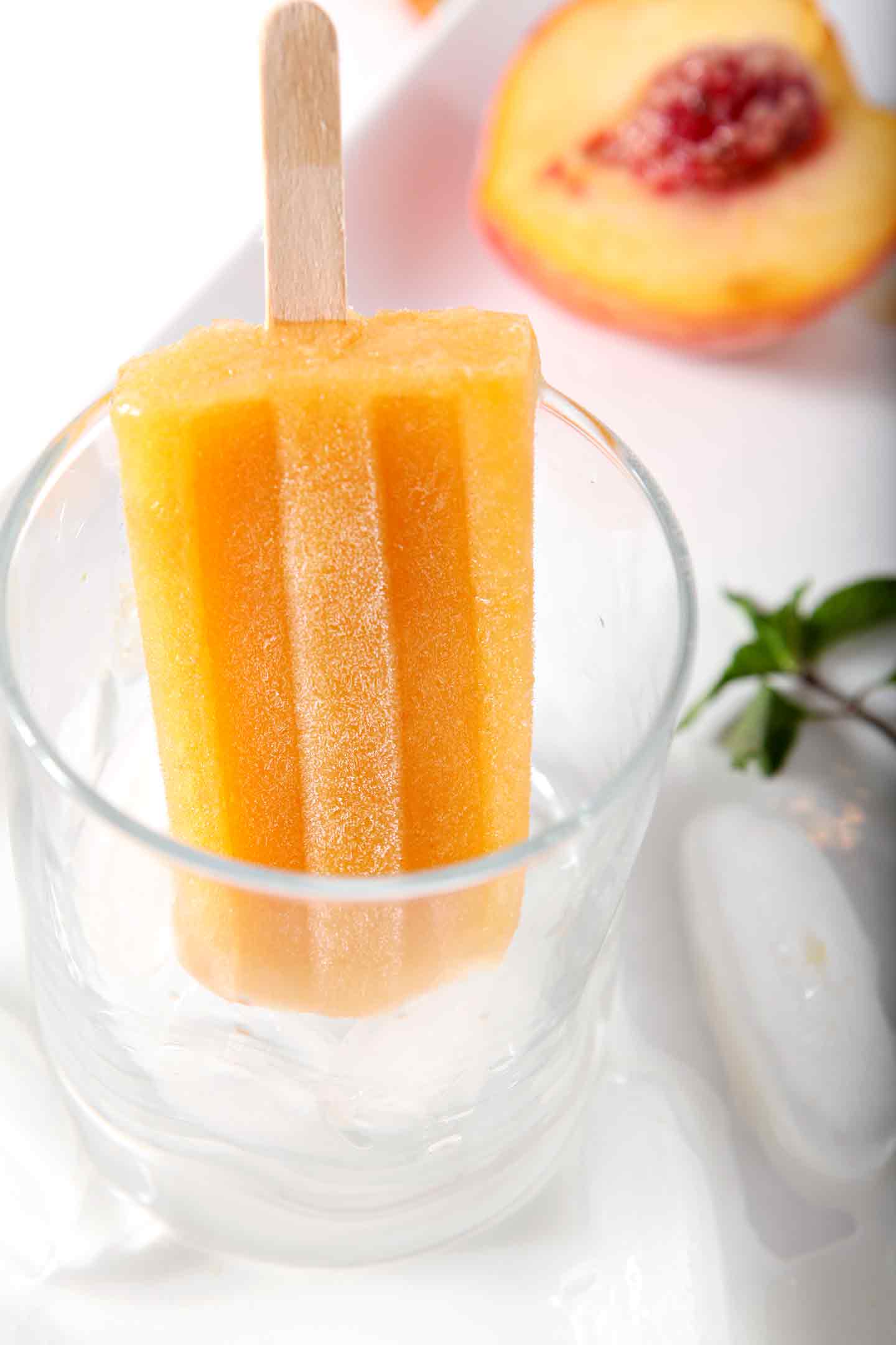 One Peach Julep Popsicle stands in a short glass, surrounded by ice cubes, peaches and mint sprigs