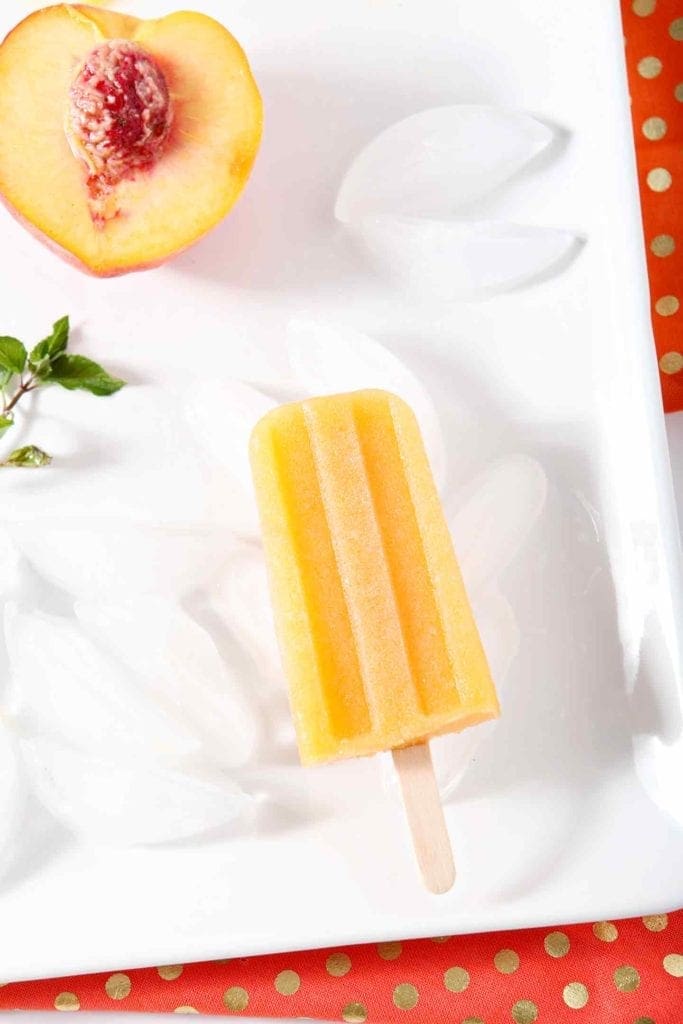 Peach Julep Popsicles are displayed on a white platter, surrounded by ice, mint sprigs and peaches