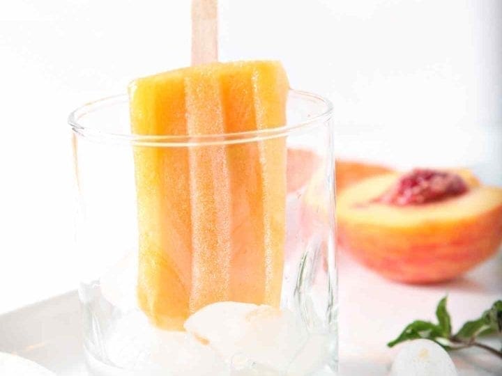 A lone Peach Julep Popsicle hangs out in a glass, surrounded by a halved peach and sprigs of mint