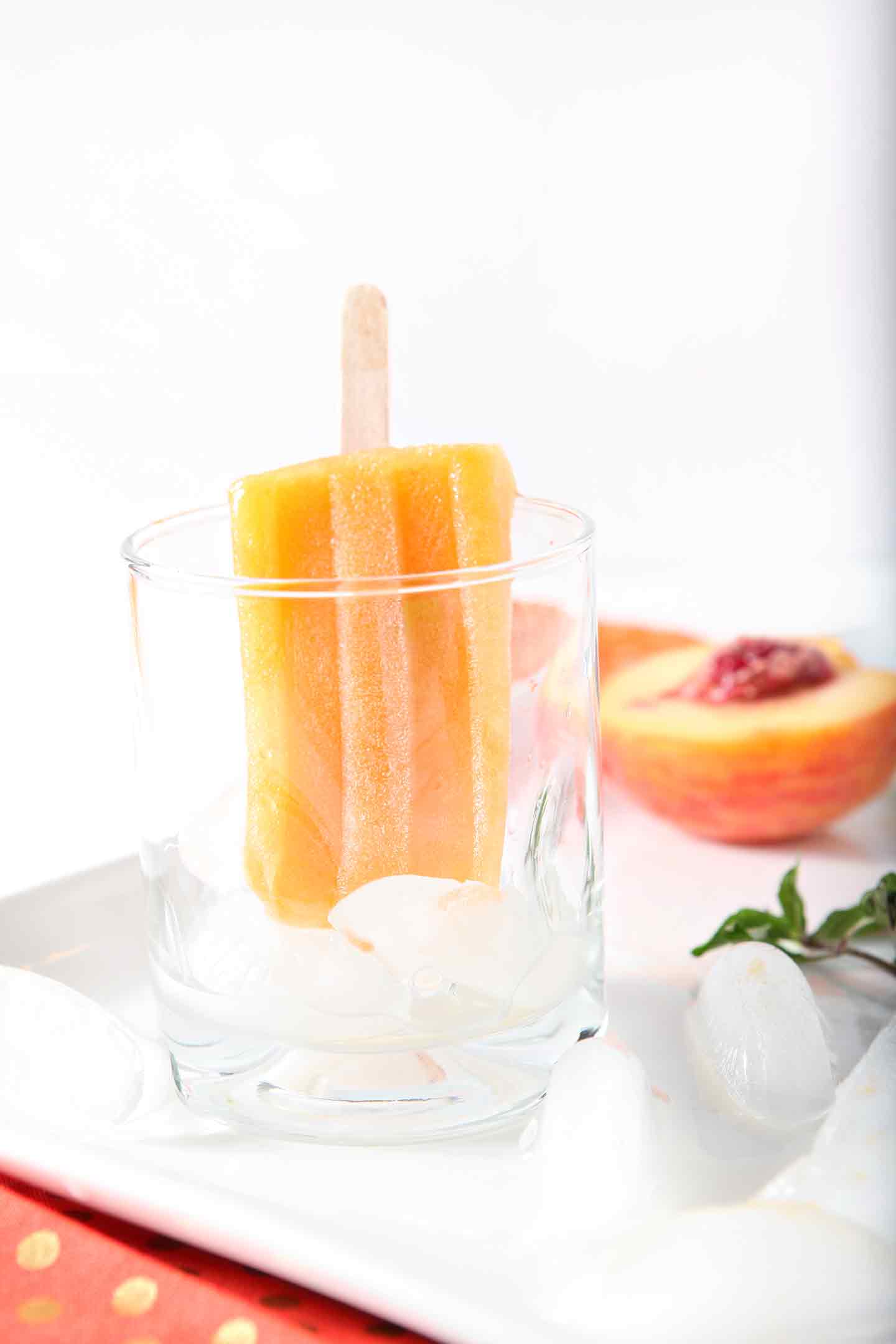 A lone Peach Julep Popsicle hangs out in a glass, surrounded by a halved peach and sprigs of mint