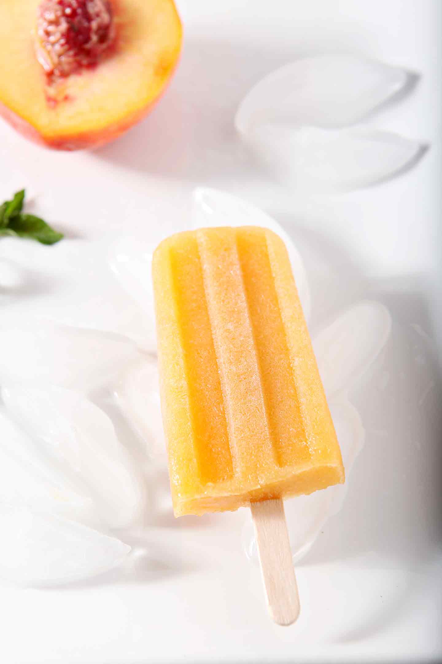 Peach Julep Popsicles are displayed on a white platter, surrounded by ice, mint sprigs and peaches