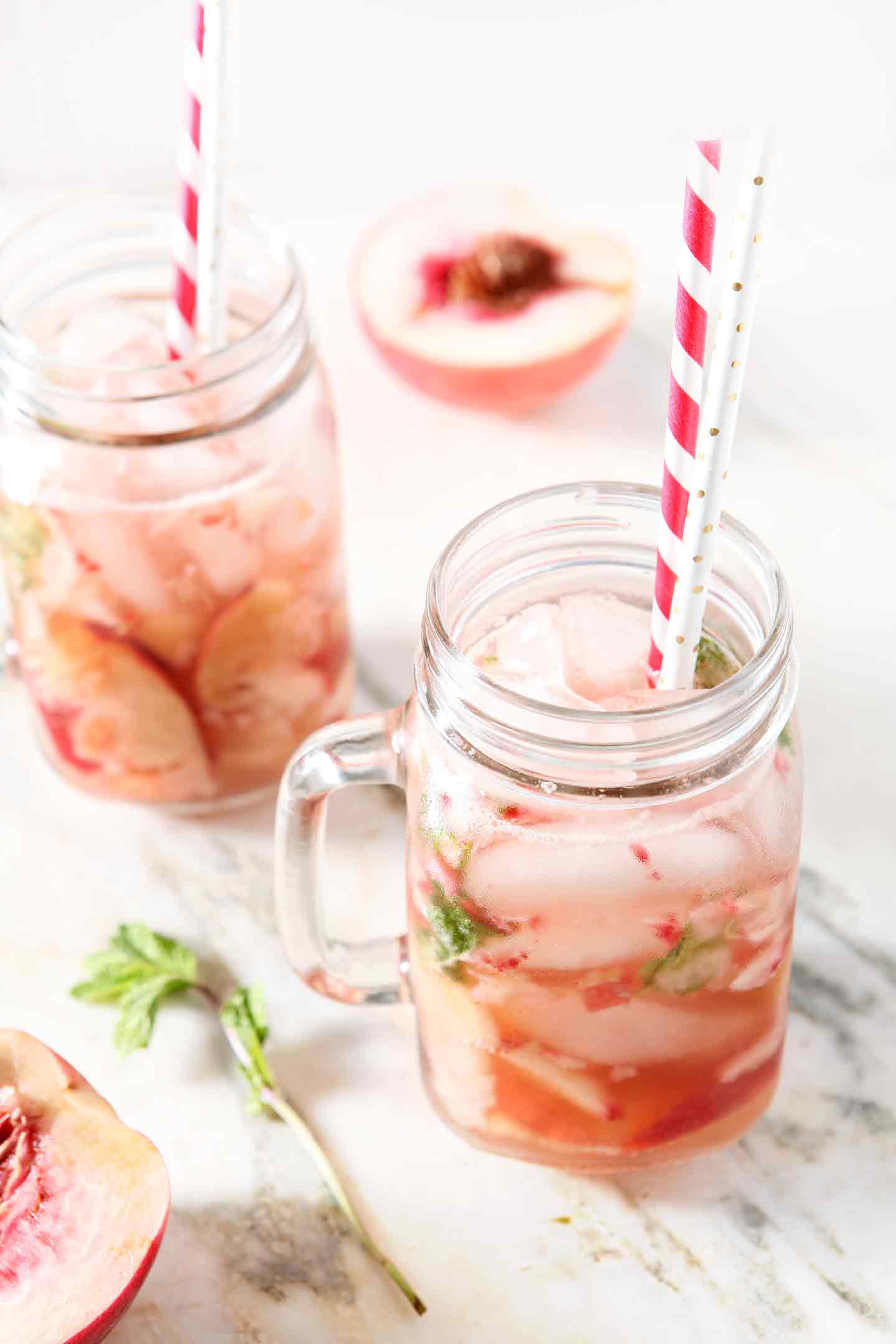 Two glasses of Ginger Peach Punch sit on a marble background, surrounded by peach halves and fresh mint.