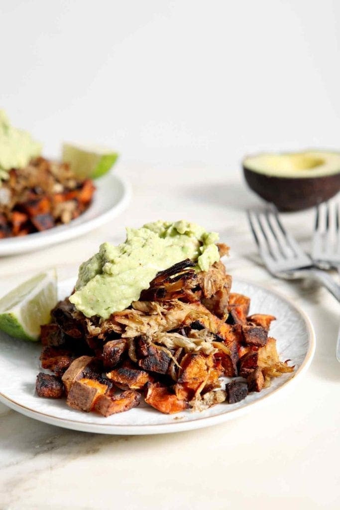 Two plates of Whole30 Slow Cooker Pork Carnitas Plates, topped with fresh guacamole and garnished with a lime slice, sit on a marble tile.