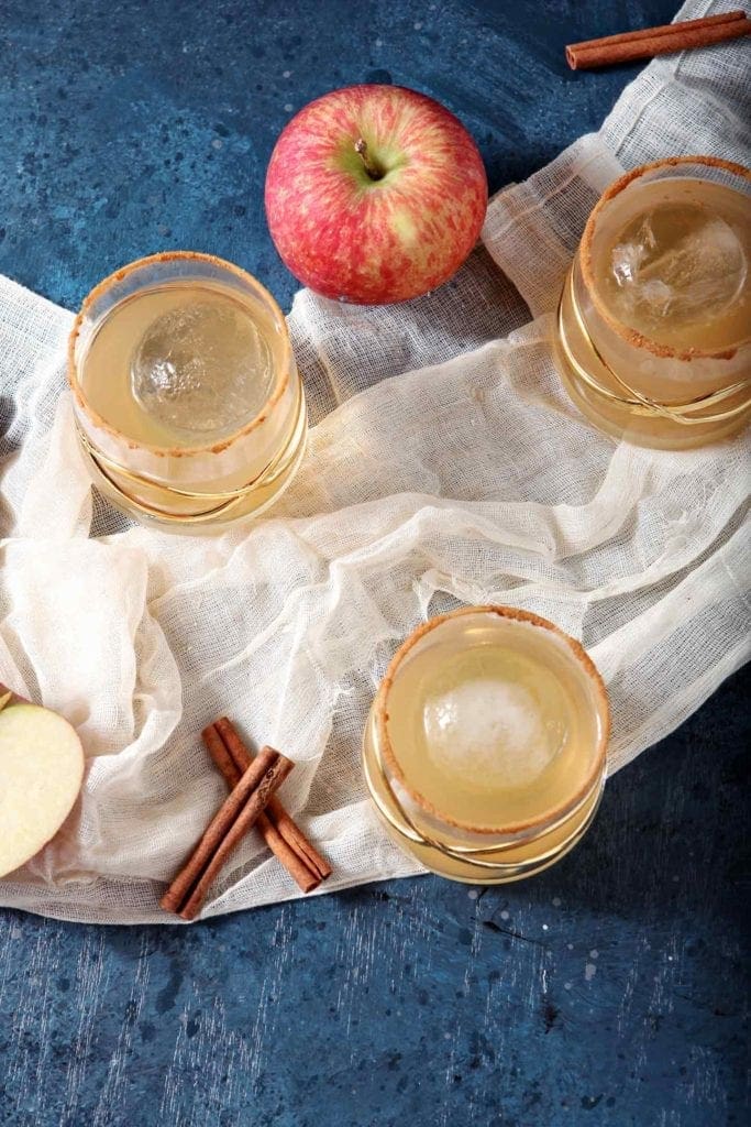 Overhead of three apple pie drinks sitting on gauzy fabric with apples and cinnamon sticks around them