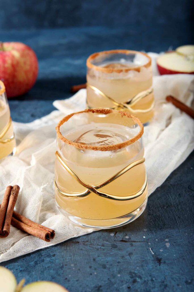 Close up of three Gin Apple Pie Drinks on a gauzy piece of fabric with apples and cinnamon sticks before being consumed