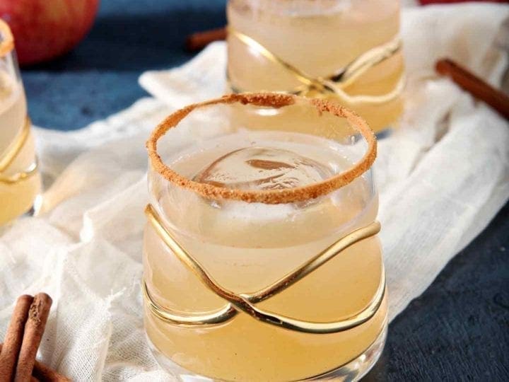 Close up of three Gin Apple Pie Drinks on a gauzy piece of fabric with apples and cinnamon sticks before being consumed