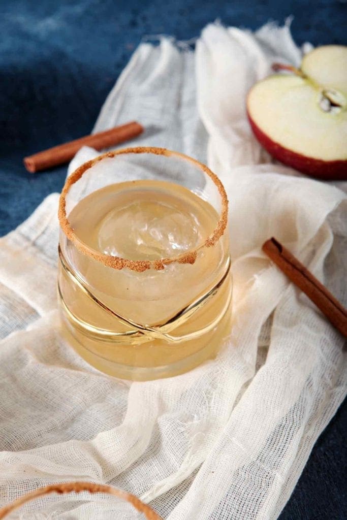 A singular glass of an apple pie drink on a gauzy white fabric surrounded with cinnamon sticks and apples