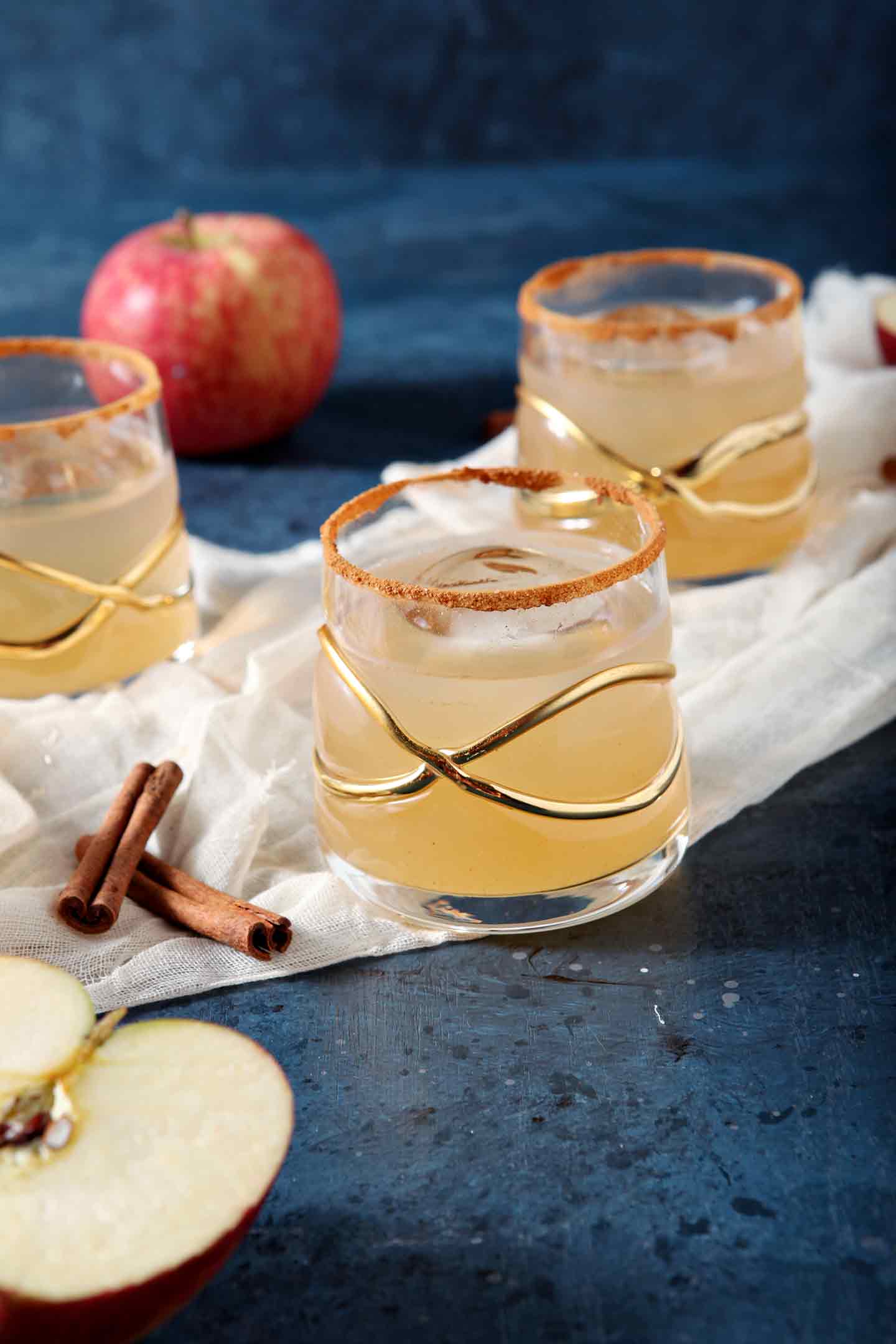 Three glasses of a Gin Apple Pie Drink sit on a gauzy fabric surrounded with apples and cinnamon sticks on a dark blue surface