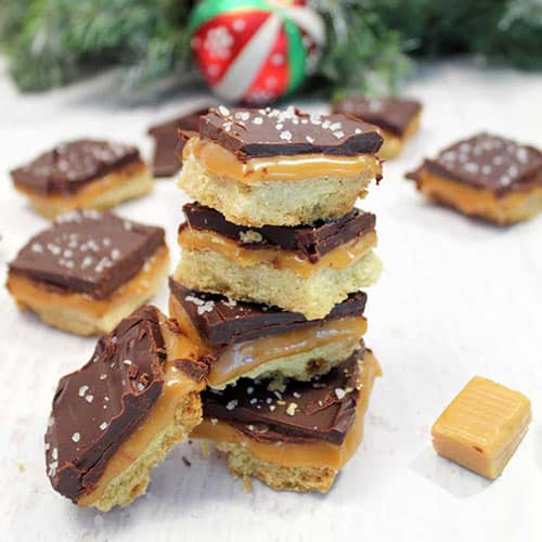 A stack of 5 shortcut millionaire bars with one bar angled off the side. Caramel candy and other bars are scattered about the white counter top.