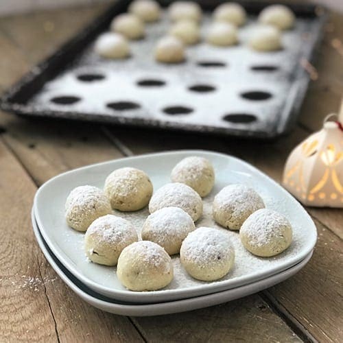 A white plate filled with polvorone cookies stacked on top of another plate with a tray of freshly powdered polvorones in the background.