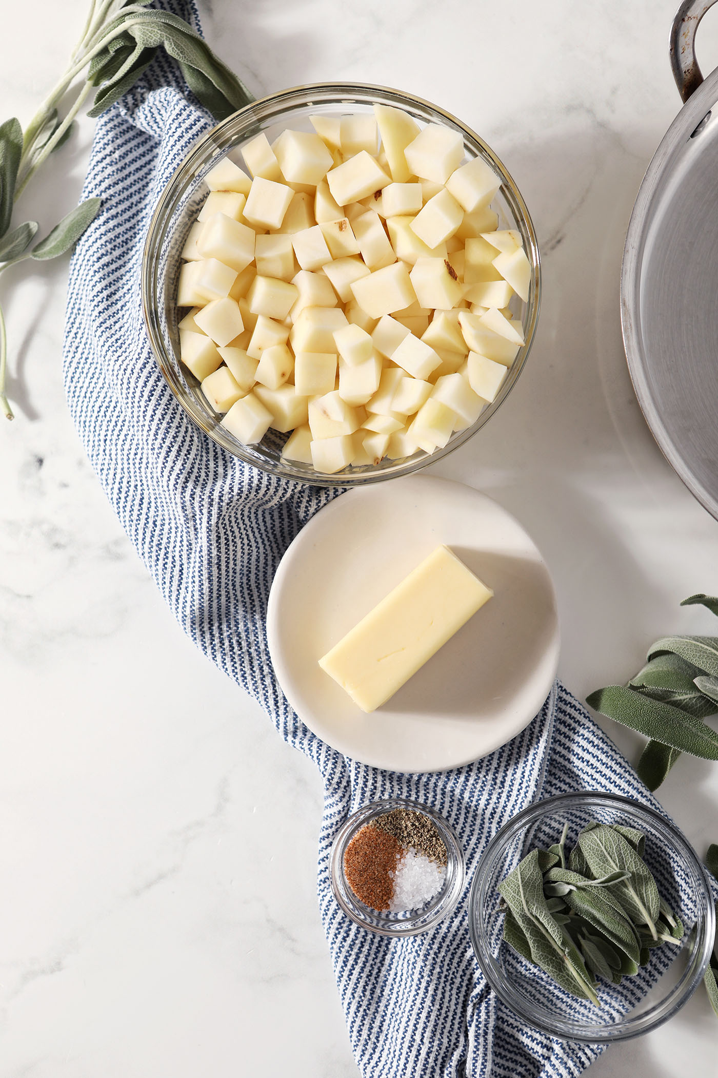 Diced potatoes in a bowl next to a platter of butter, a bowl of seasonings, and a bowl of fresh sage leaves on a marble surface with a blue-and-white-striped linen