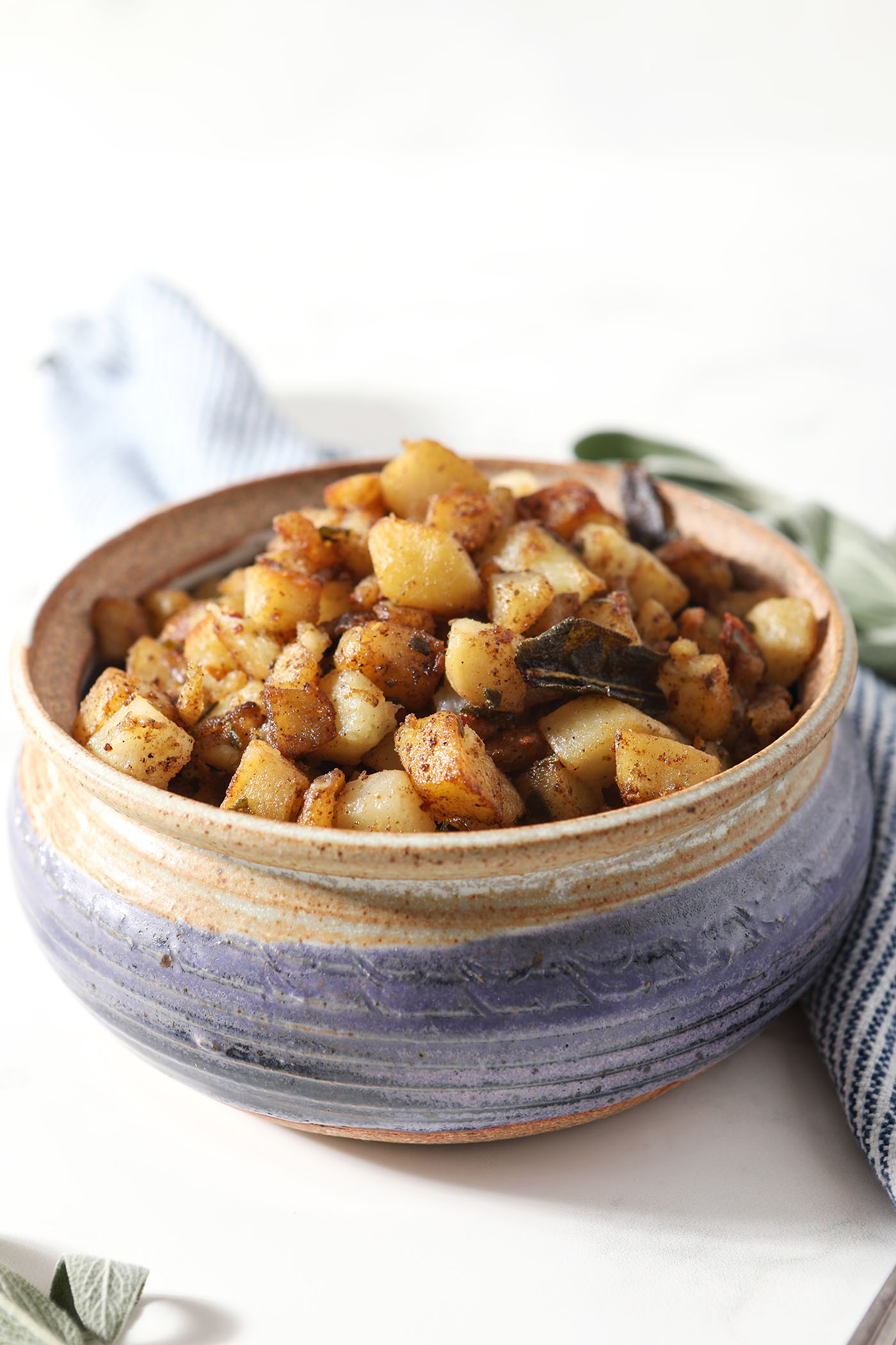 A pottery dish holding cooked home fries on a marble surface next to a blue and white linen
