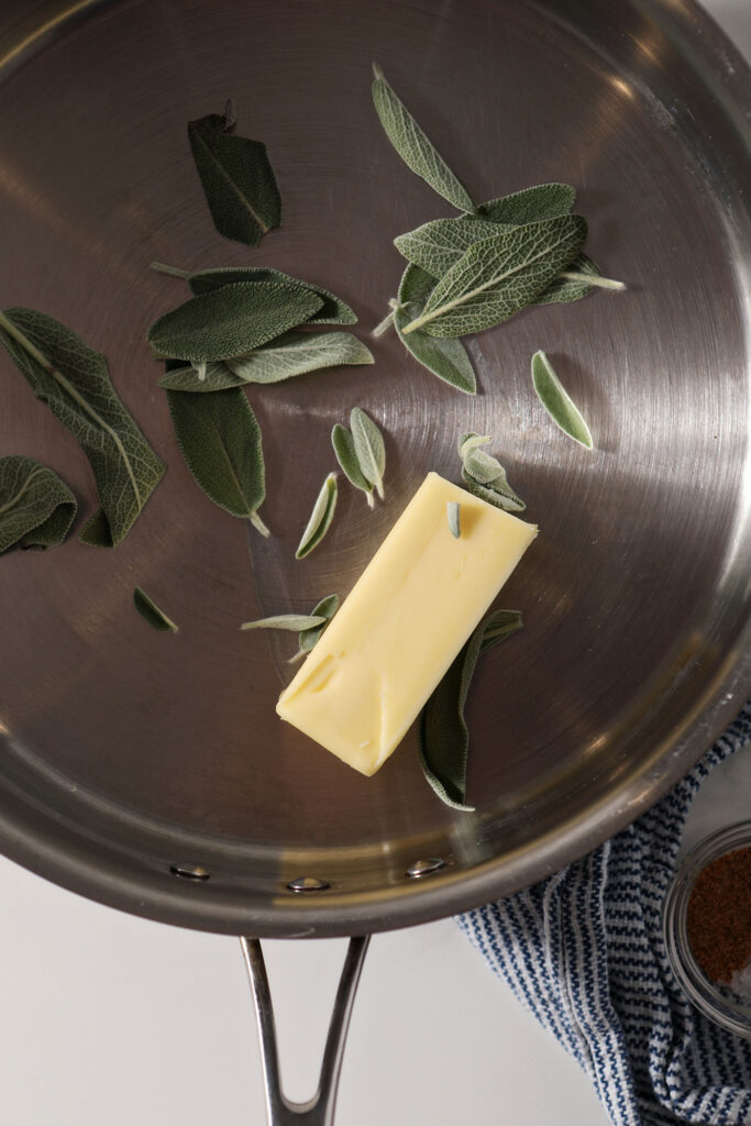 A stick of butter with fresh sage leaves in a stainless steel skillet
