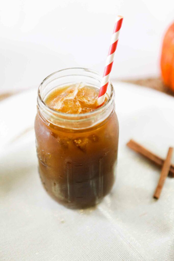 A coffee drink in a mason jar with a red and white striped straw on a white plate with cinnamon sticks around it