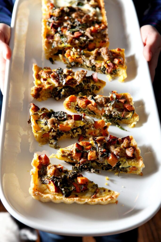 A woman holds a rectangular white tray of sausage quiche with collards and sweet potatoes in slices