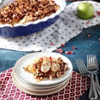 A stack of plates holds a serving of Vegan Apple Crisp with Pomegranates on a dark blue background with the serving dish behind it