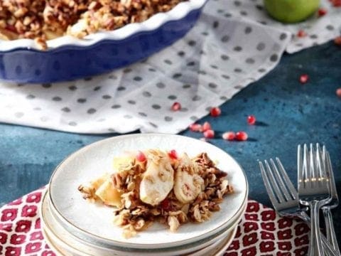 A stack of plates holds a serving of Vegan Apple Crisp with Pomegranates on a dark blue background with the serving dish behind it