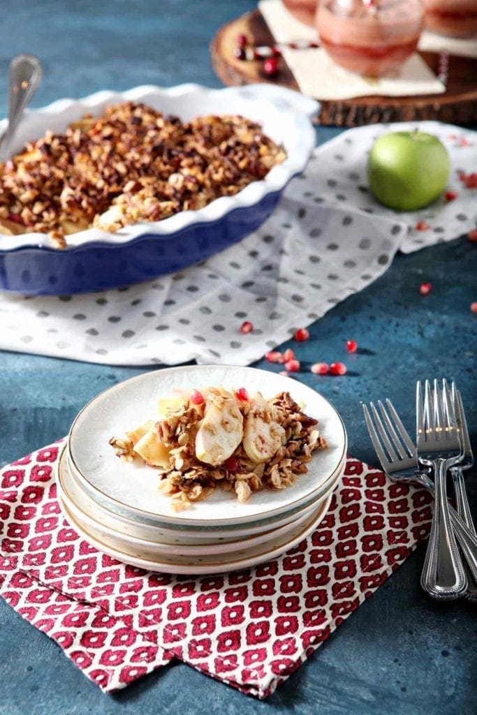 A stack of plates holds a serving of Vegan Apple Crisp with Pomegranates on a dark blue background with the serving dish behind it