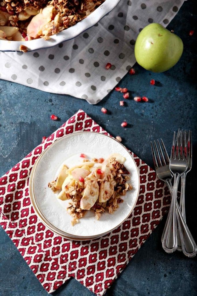 Overhead of a white plate of Vegan Apple Crisp on a patterned red napkin with apples and the baking dish on dark blue