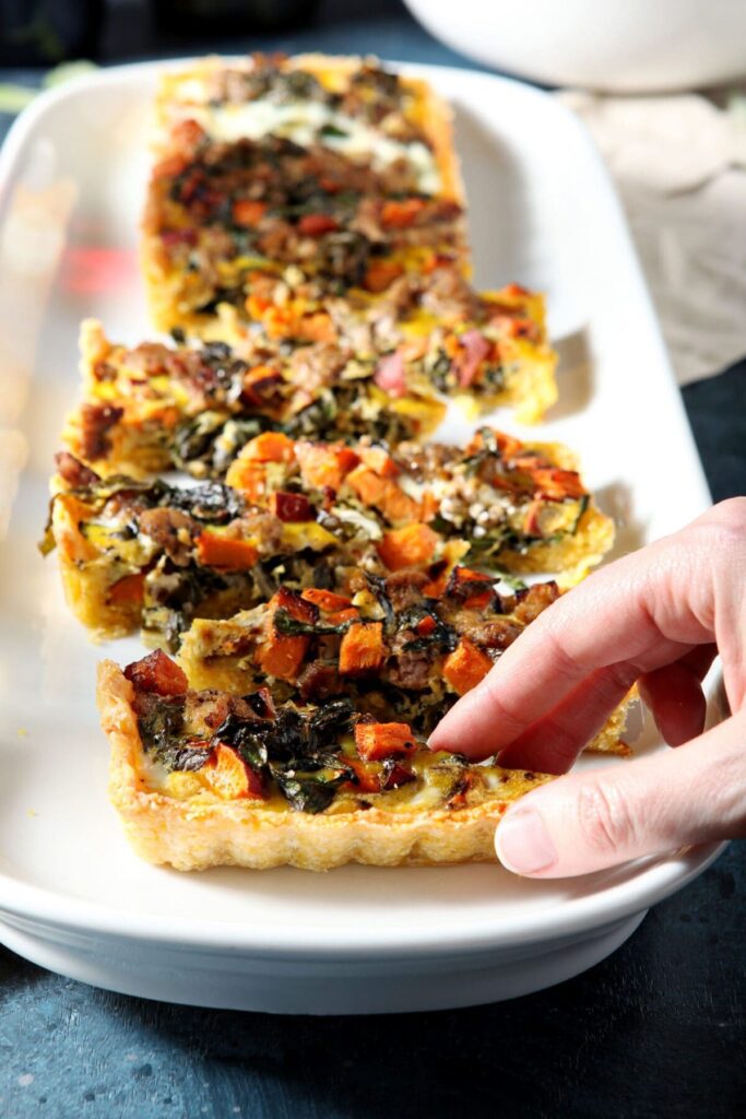 A woman grabs a bite-sized slice of a rectangular quiche from a white rectangular platter