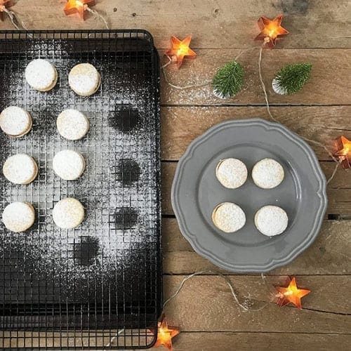 Eight Alfajore cookies on a drying rack that have just been covered in powder sugar with four cookies missing. To the right of the drying rack, is a gray plate with the four missing alfajores.