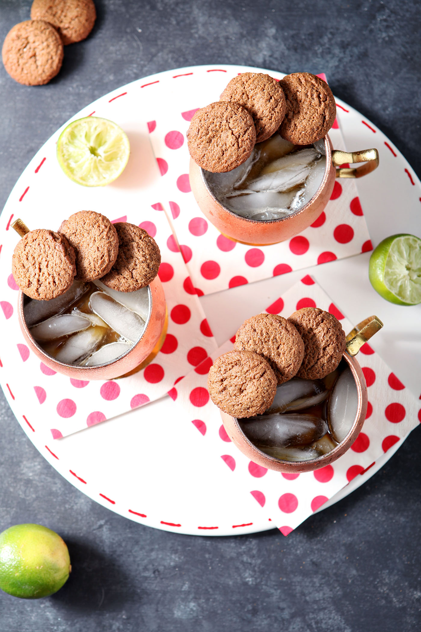 Three copper mugs holding Gingerbread Mules, garnished with ginger snaps, sit on a white platter with decorative dotted red napkins on a dark surface.