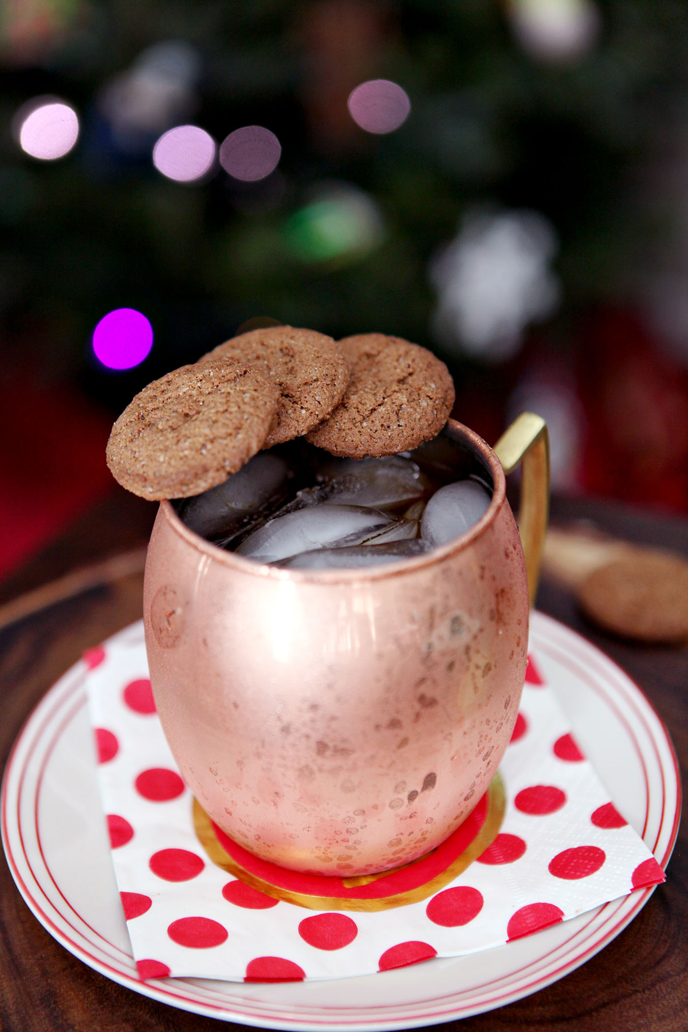 A mule and a copper mug with ginger snaps resting on the rim sit on a plate in front of a Christmas tree.
