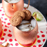 Two copper mugs of drinks, garnished with three gingersnaps on the rim, sit on a white tray with decorative red and white napkins.