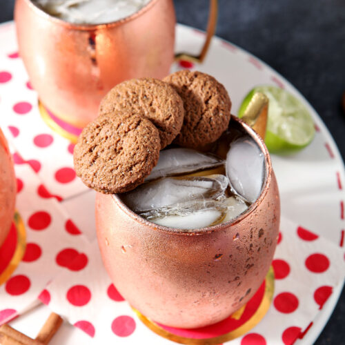 Two copper mugs of drinks, garnished with three gingersnaps on the rim, sit on a white tray with decorative red and white napkins.