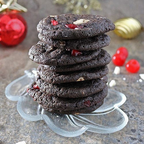 A vintage glass leaf plate holding a vertical stack of chocolate peppermint cherry cookies with cherries and chocolate scattered around the plate.