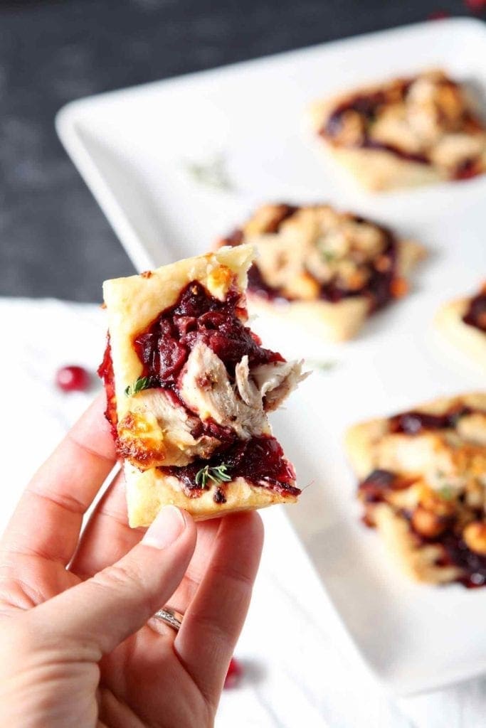 A woman holds a half-eaten Cranberry Turkey Puff Pastry Tartlet next to a serving plate with more of the tartlets