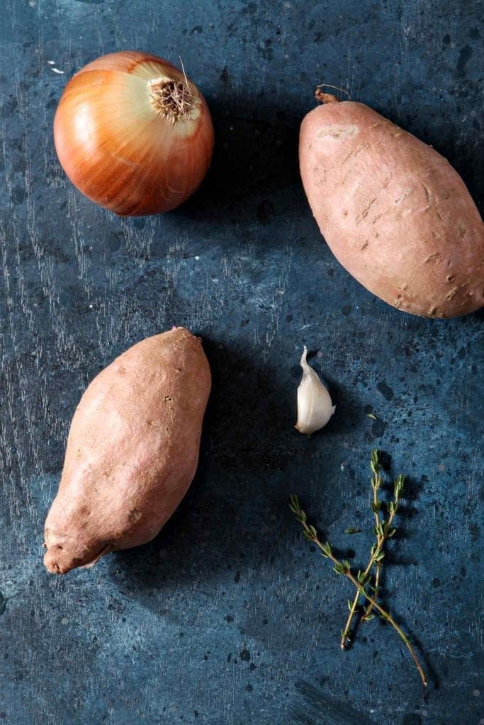 Two sweet potatoes, a clove of garlic, fresh thyme and an onion sit together on a dark countertop
