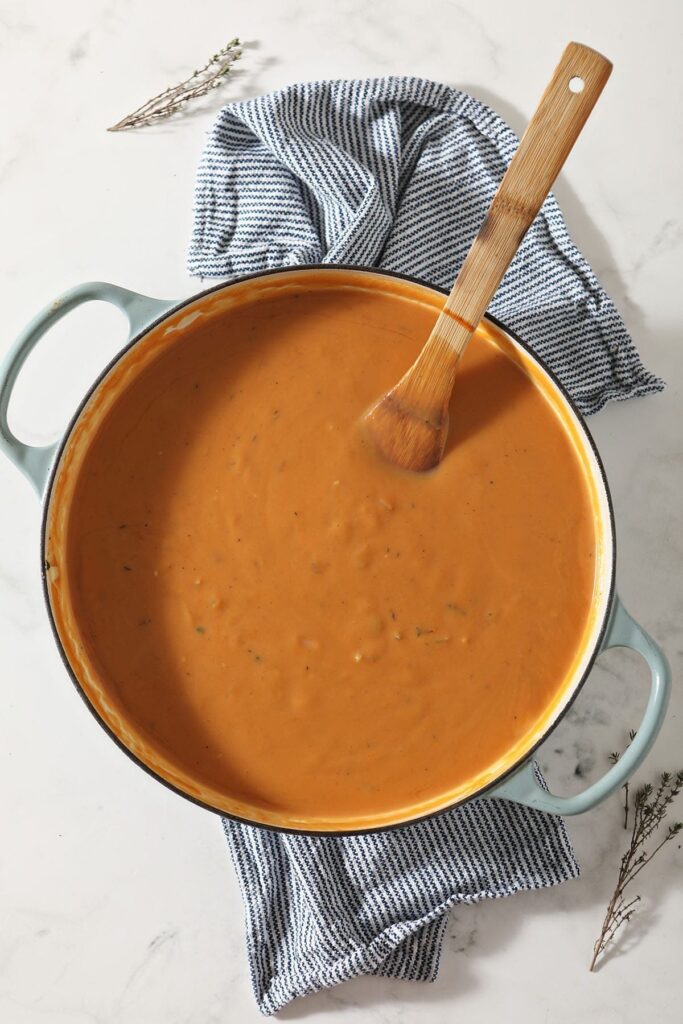 A saucepan holding Sweet Potato Bisque with a wooden spoon on it sits on a blue striped towel on a marble counter