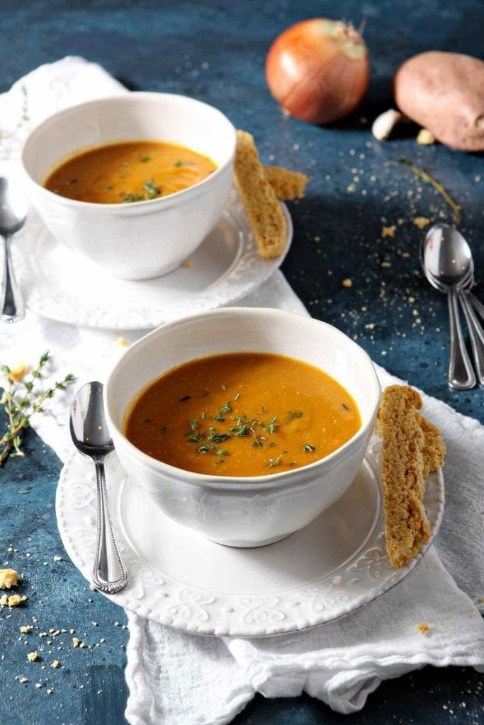 Two bowls of Sweet Potato Soup on a blue background with cornbread biscotti