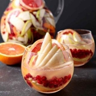 Close up of a glass holding Winter White Sangria, garnished with fresh fruit and in front of another glass and a pitcher on a dark countertop