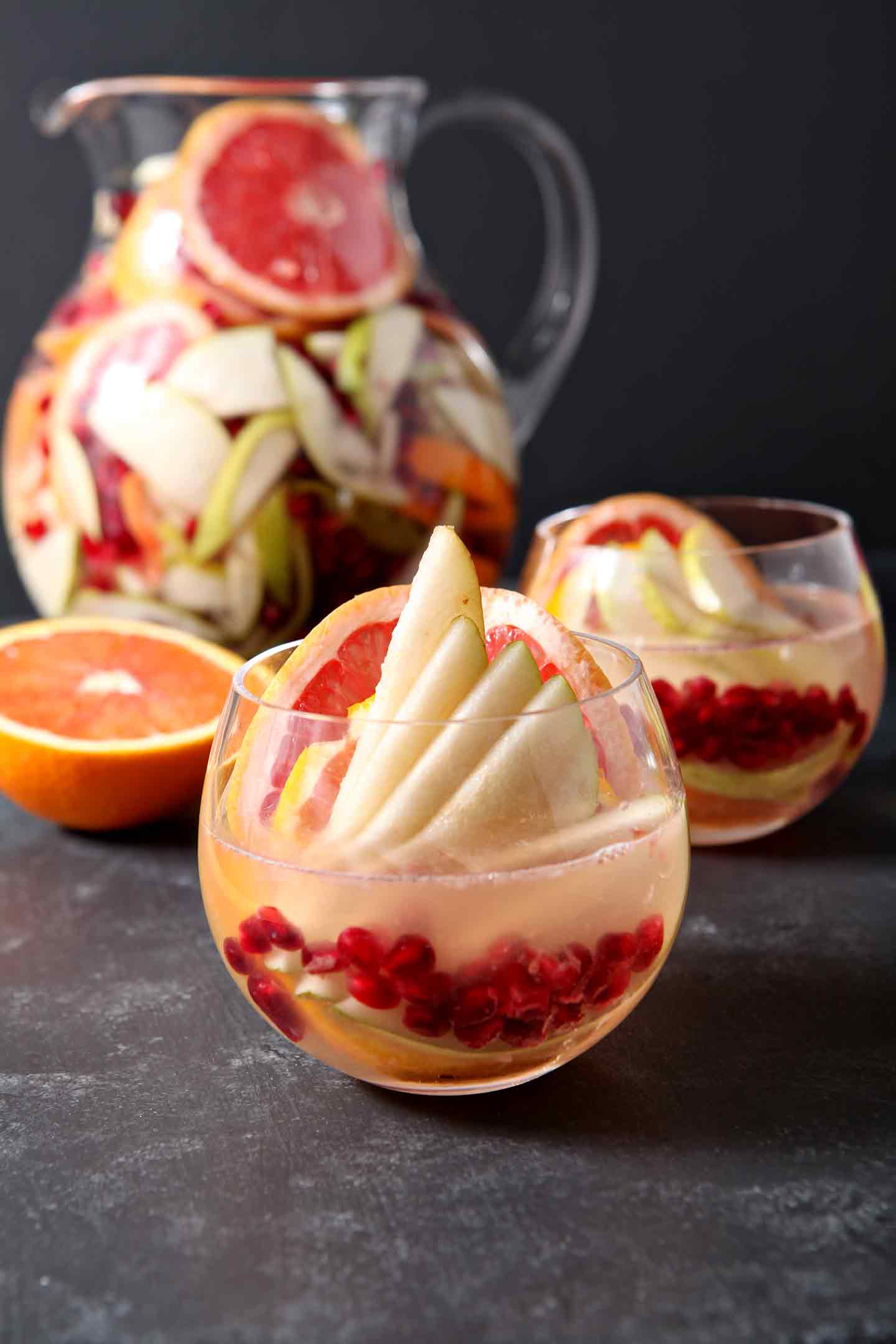 Close up of a glass holding Winter White Sangria, garnished with fresh fruit and in front of another glass and a pitcher on a dark countertop