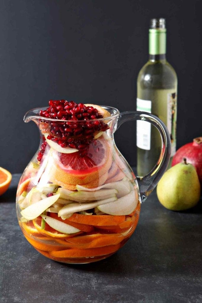 A pitcher holds oranges, pears, grapefruit and pomegranate arils on a dark surface