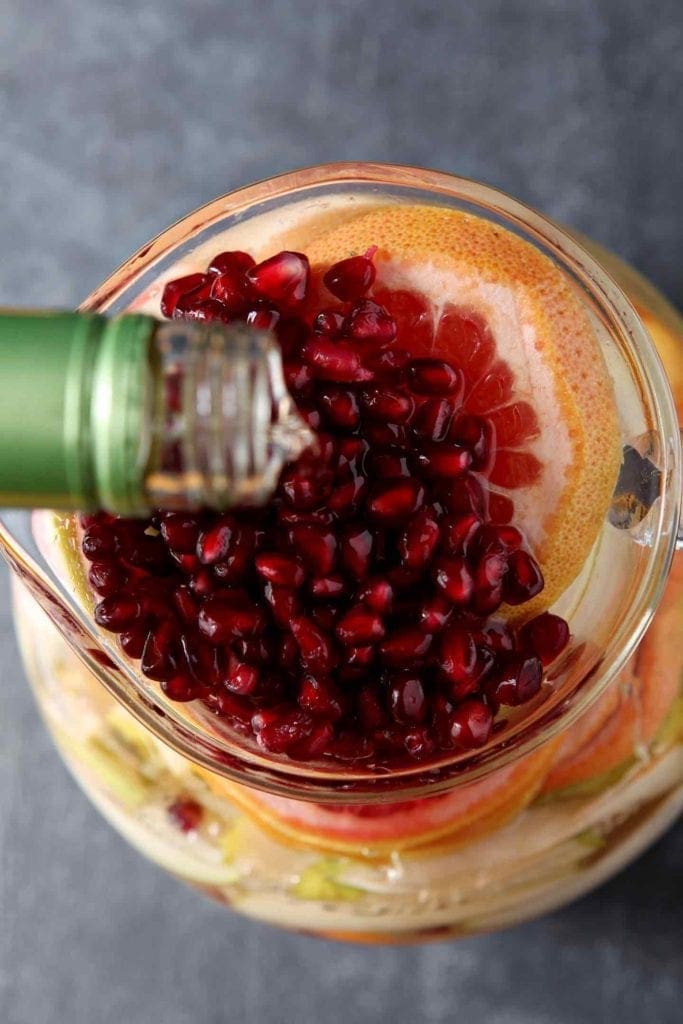 White wine pours on top of pomegranate arils and grapegruit rounds in a pitcher