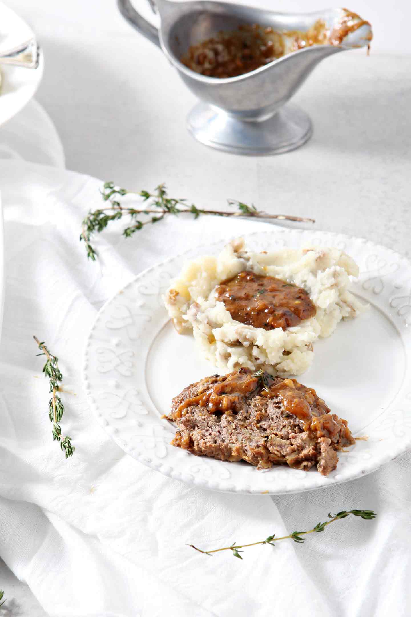 A slice of French Onion Meatloaf, covered in gravy, on a white plate with a serving of mashed potatoes and gravy, ready for eating