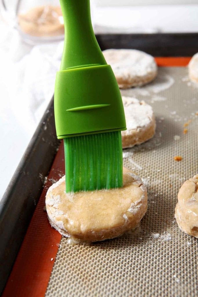 A green pastry brush brushes butter onto a biscuit on a baking sheet