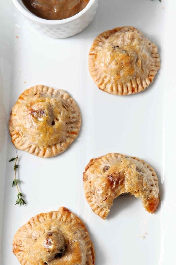 Several meat hand pies with mashed potatoes on a white platter next to a bowl of gravy