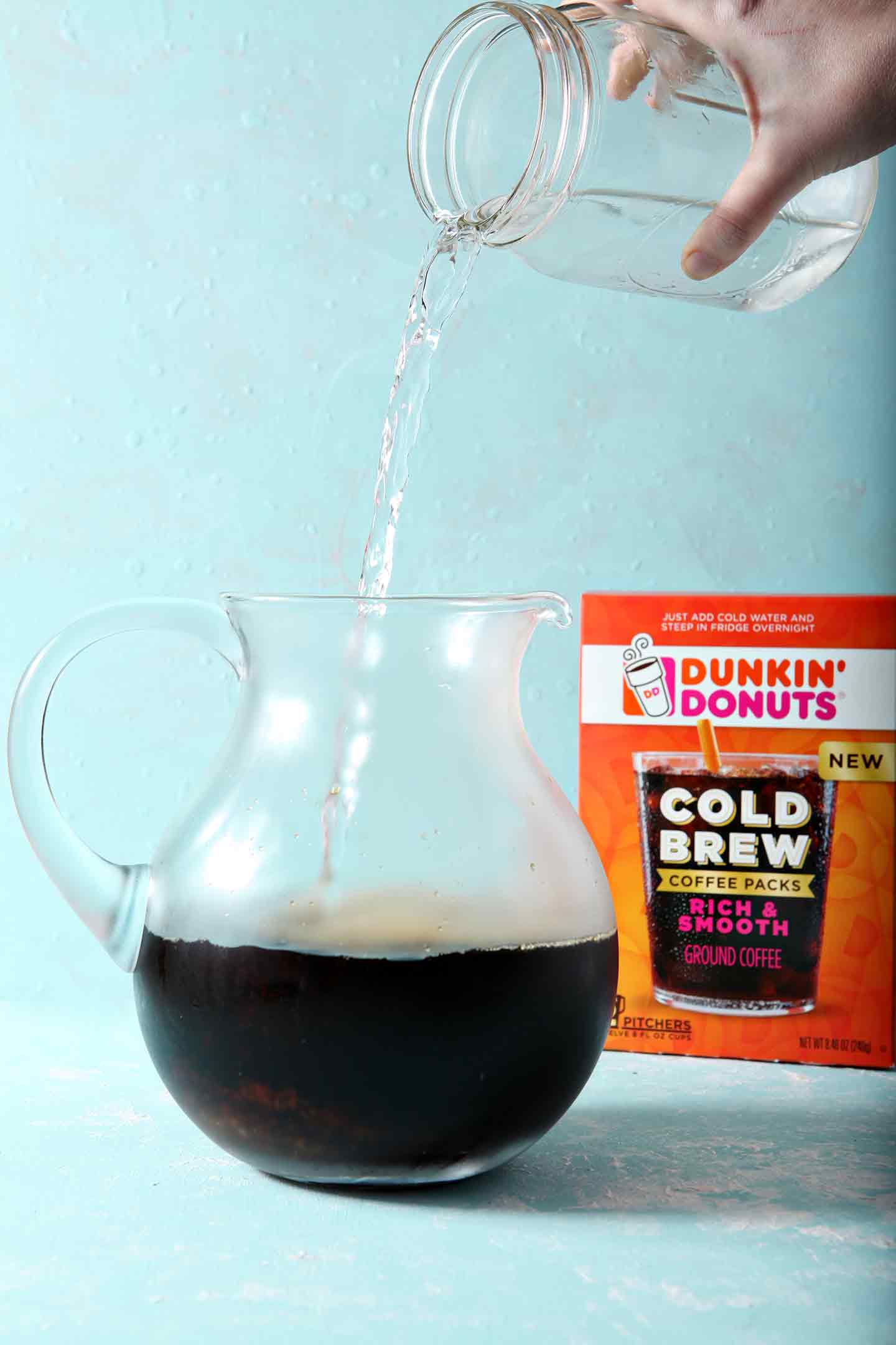 Water being poured on top of Dunkin' Donuts Cold Brew packets, on a turquoise background, before going in the fridge to steep overnight